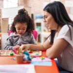 Teacher and toddler playing with maths puzzle game sitting on table at kindergarten